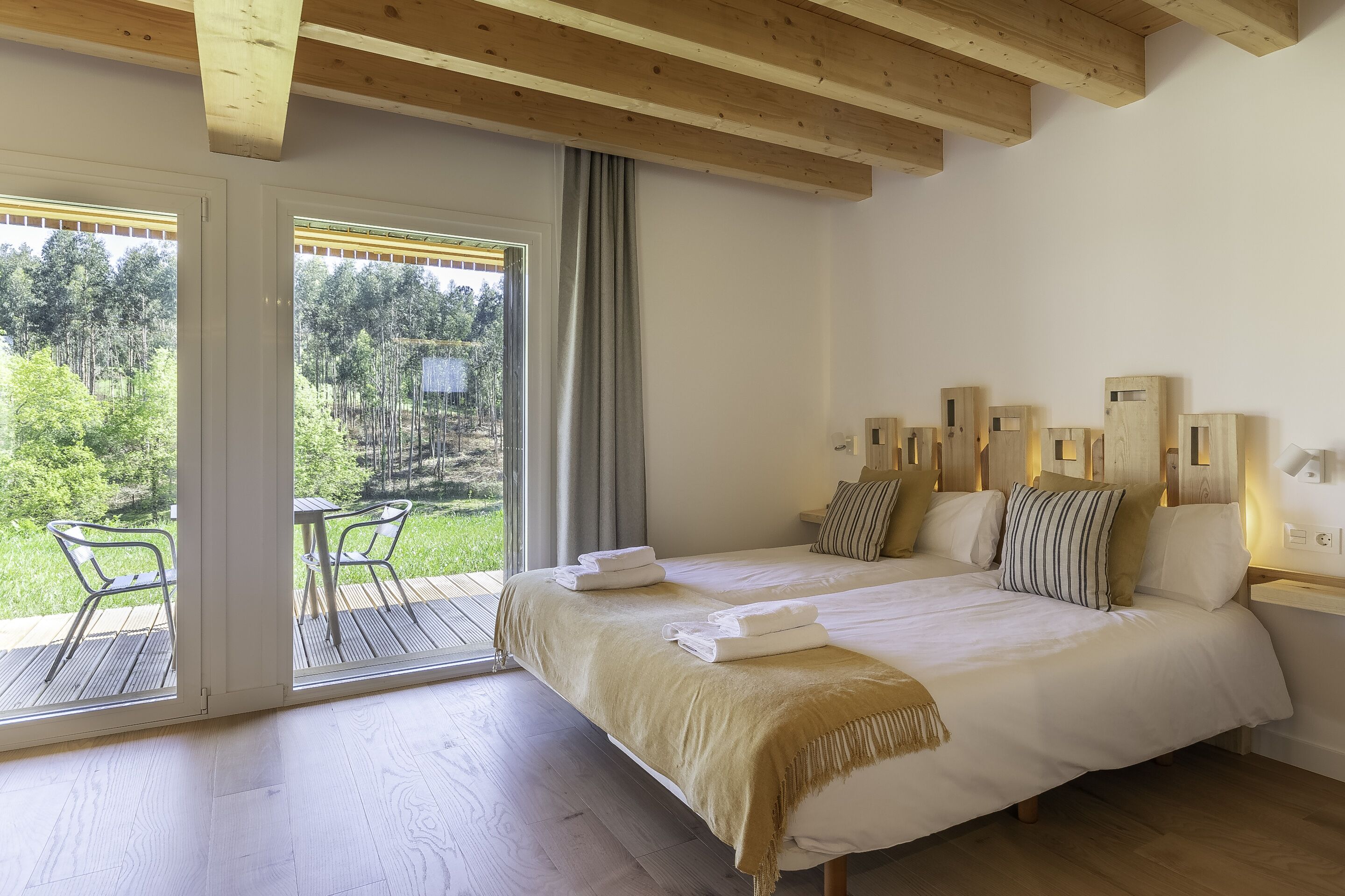 Un dormitorio con una cama grande, techo de madera, vista a la vegetación por una puerta corrediza y una pequeña mesa con sillas.