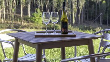 Una mesa con dos copas de vino y una botella, ubicada al aire libre con sillas metálicas y un bosque de fondo.