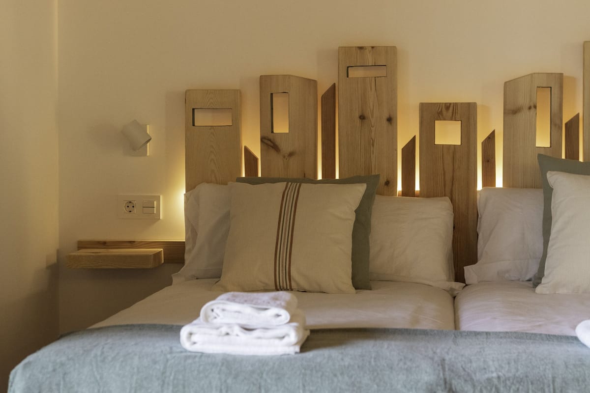 Quarto de hotel com cama, almofadas, mesa de cabeceira e luminária fixa na parede.