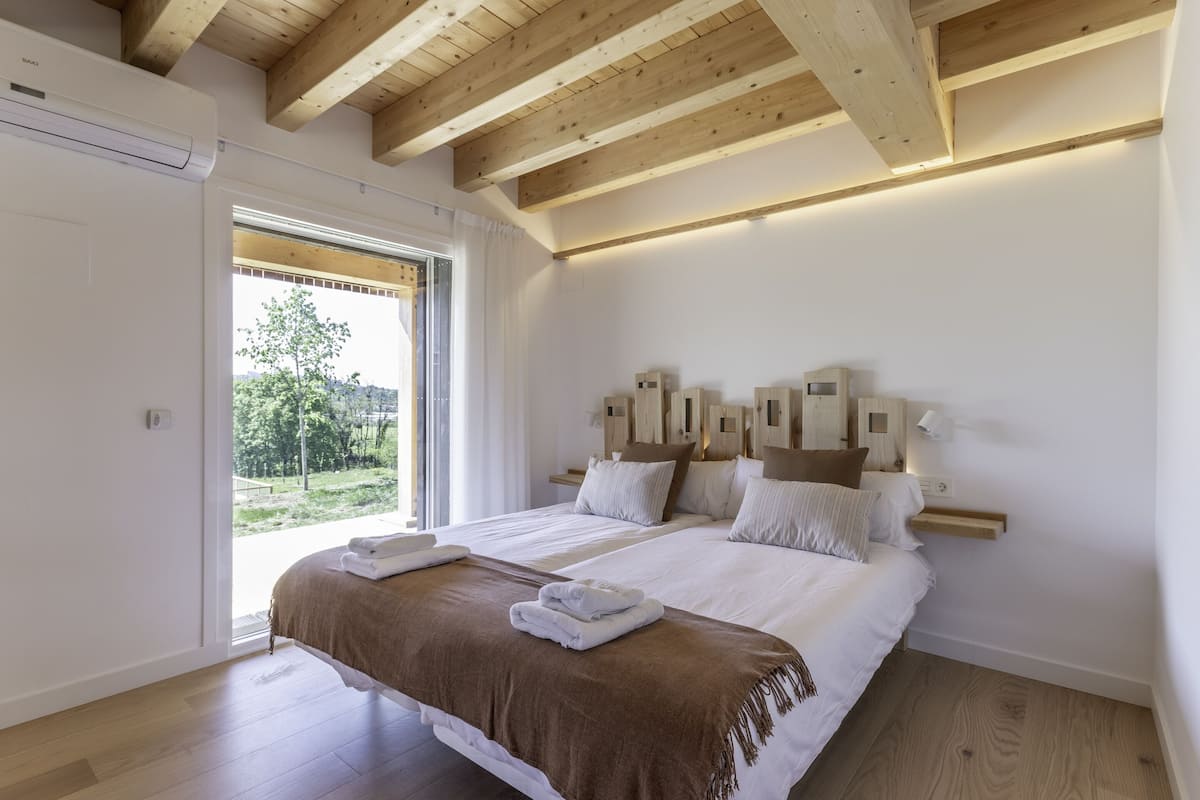 Quarto com cama grande, vigas de madeira no teto, mesa de cabeceira com abajur e vista para o verde lá fora.