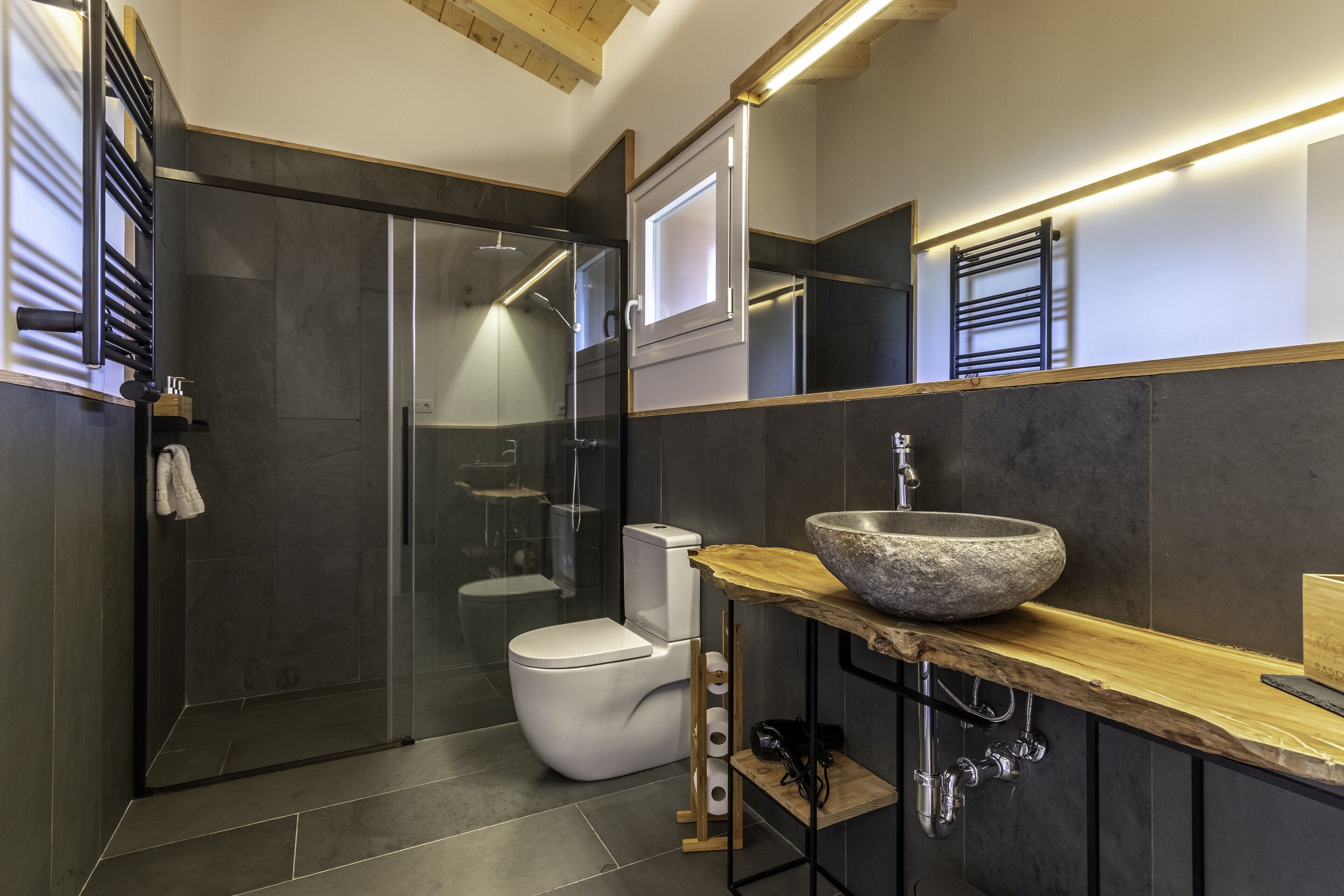 A modern bathroom with a glass-enclosed shower, a white toilet, and a large stone sink on a wooden countertop.