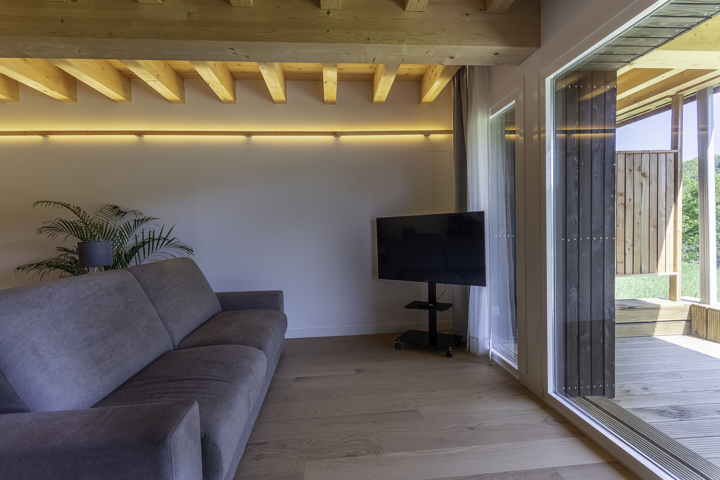 Una sala de estar moderna con un sofá gris, un televisor de pantalla plana sobre un mueble bajo, y un amplio ventanal corredizo que da a una terraza exterior.