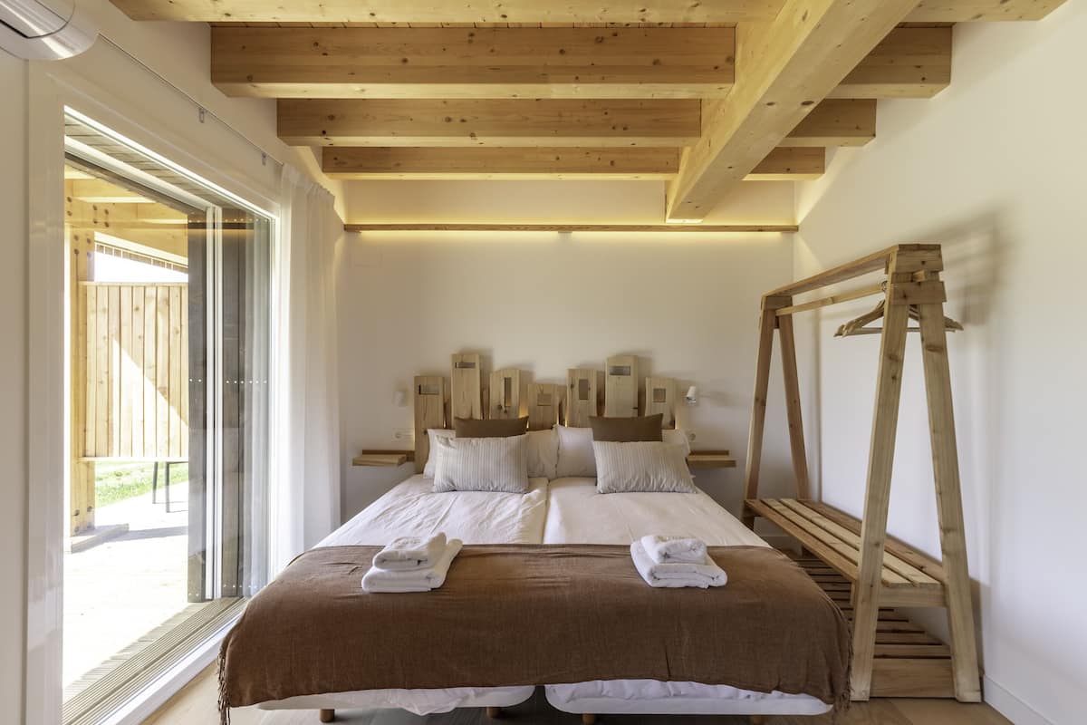 Quarto com teto de madeira, cama com roupa de cama branca, escada de madeira e uma janela.