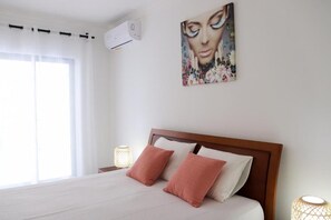 Une chambre d’hôtel avec un grand lit, du linge de lit blanc, deux oreillers roses, une tête de lit en bois, un climatiseur fixé au mur, une fenêtre avec des rideaux légers et deux lampes de chevet.