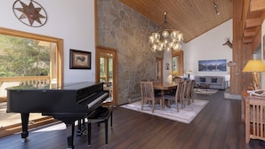 A spacious living room with a grand piano, a stone wall, wooden ceiling, and a large dining table. - Alpenglow Retreat | Big Sky Town | Sleeps 12! (Big Sky)