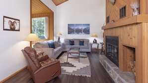 A cozy living room with a fireplace, a sofa, a leather chair, and a large window. - Alpenglow Retreat | Big Sky Town | Sleeps 12! (Big Sky)