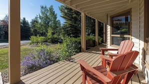 A wooden deck with Adirondack chairs, a glass door, and a view of a garden with trees and shrubs. - Alpenglow Retreat | Big Sky Town | Sleeps 12! (Big Sky)