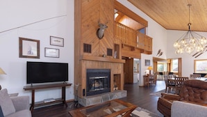 A spacious living room with a fireplace, wooden walls, a large TV, and a comfortable seating area. - Alpenglow Retreat | Big Sky Town | Sleeps 12! (Big Sky)