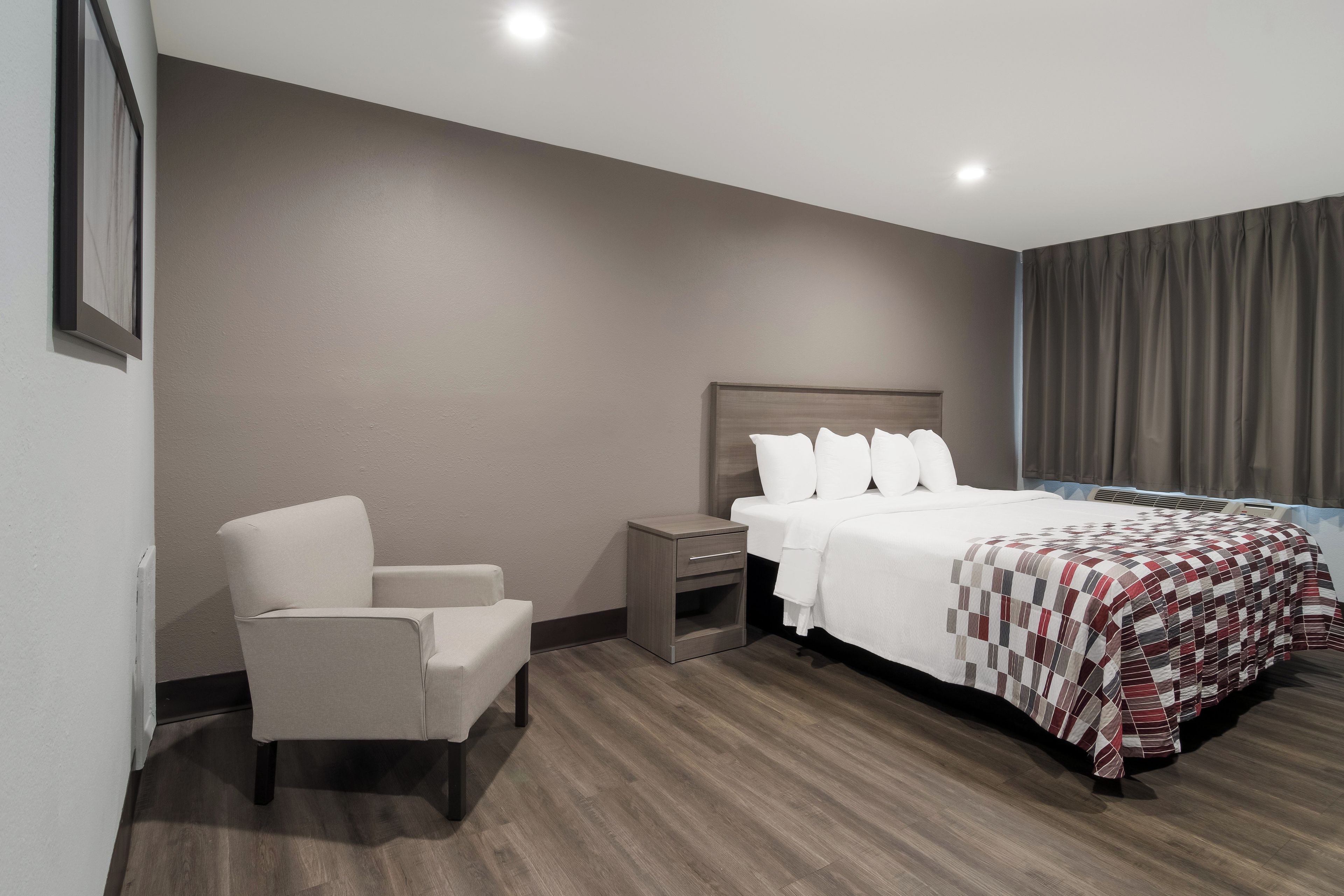 Una habitación de hotel con cama, una silla, una mesita de noche y un gran ventanal con cortinas.