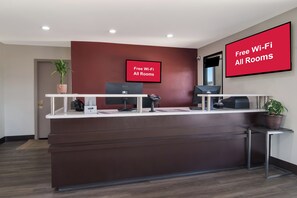 A reception desk with a red wall, two monitors, a phone, and a sign stating 'Free Wi-Fi All Rooms'. - Red Roof Inn Rocky Mount - Battleboro (Battleboro)