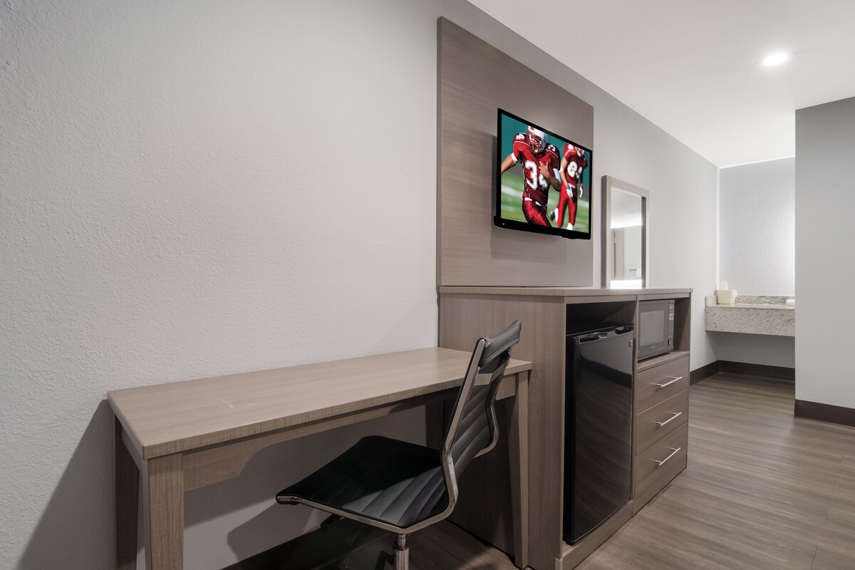 A modern hotel room with a wooden desk, a chair, a flat-screen TV mounted on the wall, a microwave, and a small kitchenette area.