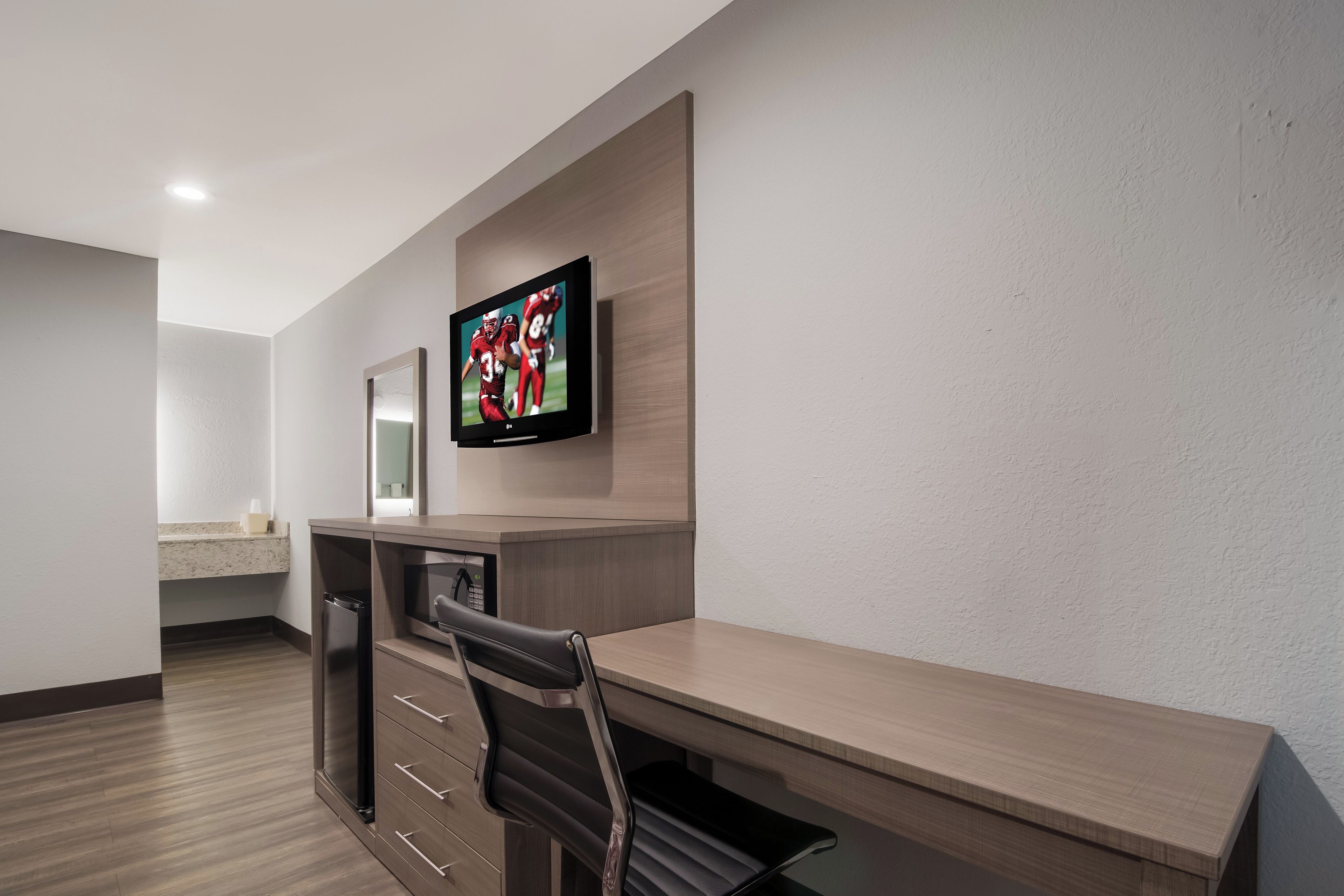 A hotel room with a wooden desk, a television mounted on the wall, a microwave, and a small kitchenette area.