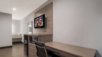 A hotel room with a wooden desk, a television mounted on the wall, a microwave, and a small kitchenette area.