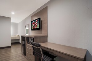 A hotel room with a wooden desk, a television mounted on the wall, a microwave, and a small kitchenette area.