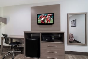 A hotel room with a flat-screen TV mounted on the wall, a desk with a chair, a microwave, and a drawer unit.
