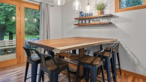 Un coin repas avec une table en bois et des chaises en métal, une étagère ornée d’objets décoratifs et une fenêtre donnant sur un paysage verdoyant.