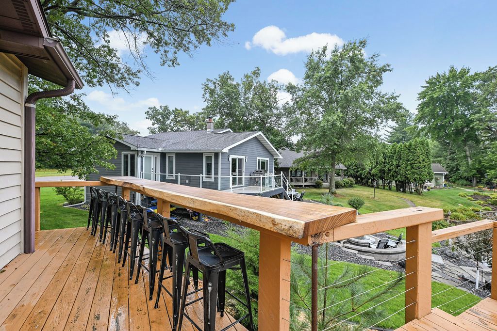 Una terraza con taburetes de bar y barandilla de madera, con vista a un jardín ajardinado y una casa al fondo.