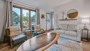 Un salon avec une grande table en bois, un canapé et un fauteuil. Il y a une fenêtre avec des rideaux, une porte et une étagère fixée au mur, ornée d’un miroir et d’objets décoratifs.