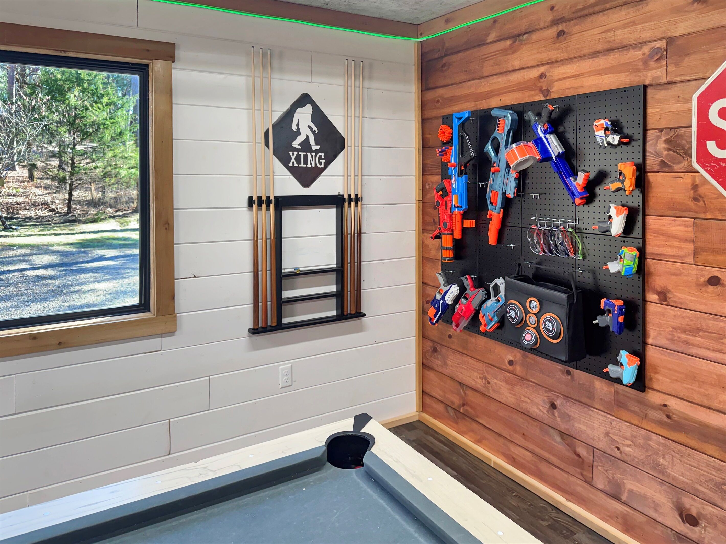 Een kamer met een tafeltennistafel, een houten wand, een prikbord vol speelgoed en een raam met uitzicht op bomen.