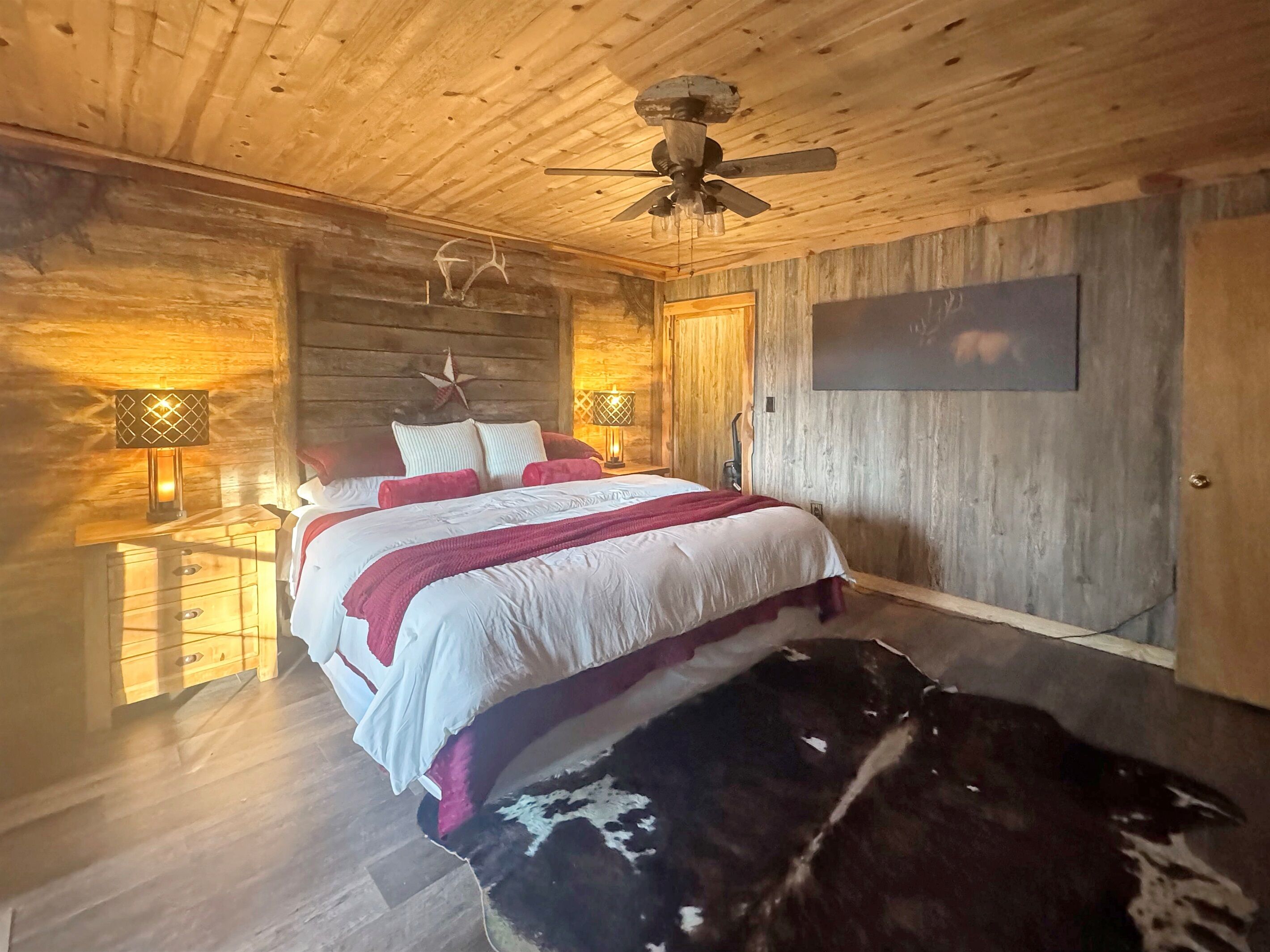 A wooden cabin-style bedroom with a bed, two bedside lamps, a ceiling fan, and a mounted picture.
