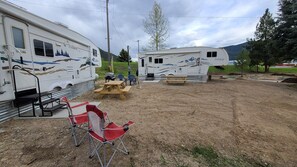 Un camping con dos vehículos recreativos, mesas de picnic y una silla plegable roja.