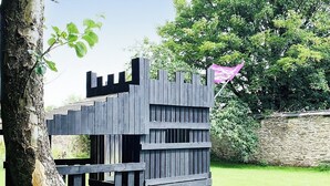 A wooden playhouse with a ramp entrance, surrounded by a stone wall and greenery.
