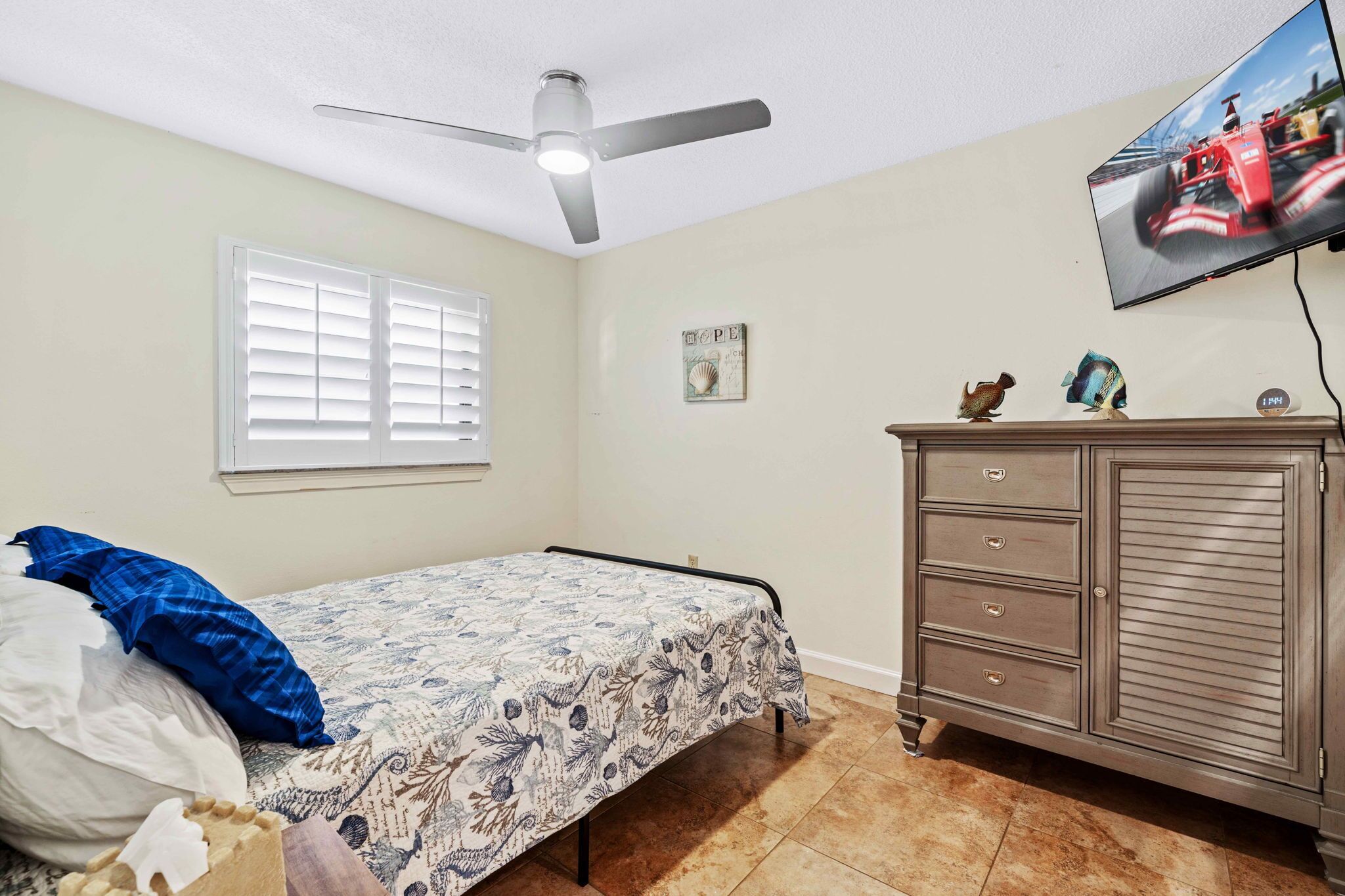 Un dormitorio con cama, ventilador de techo, televisor, cómoda y ventana con persianas.