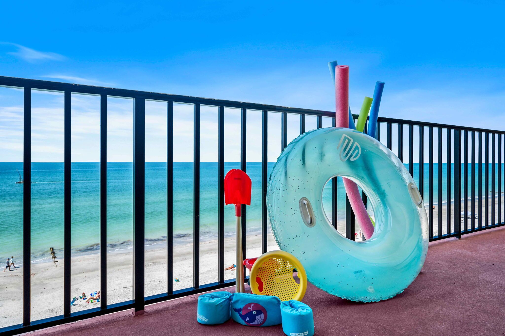A balcony with a view of the beach, featuring a large inflatable ring, a red shovel, and a yellow paddle.