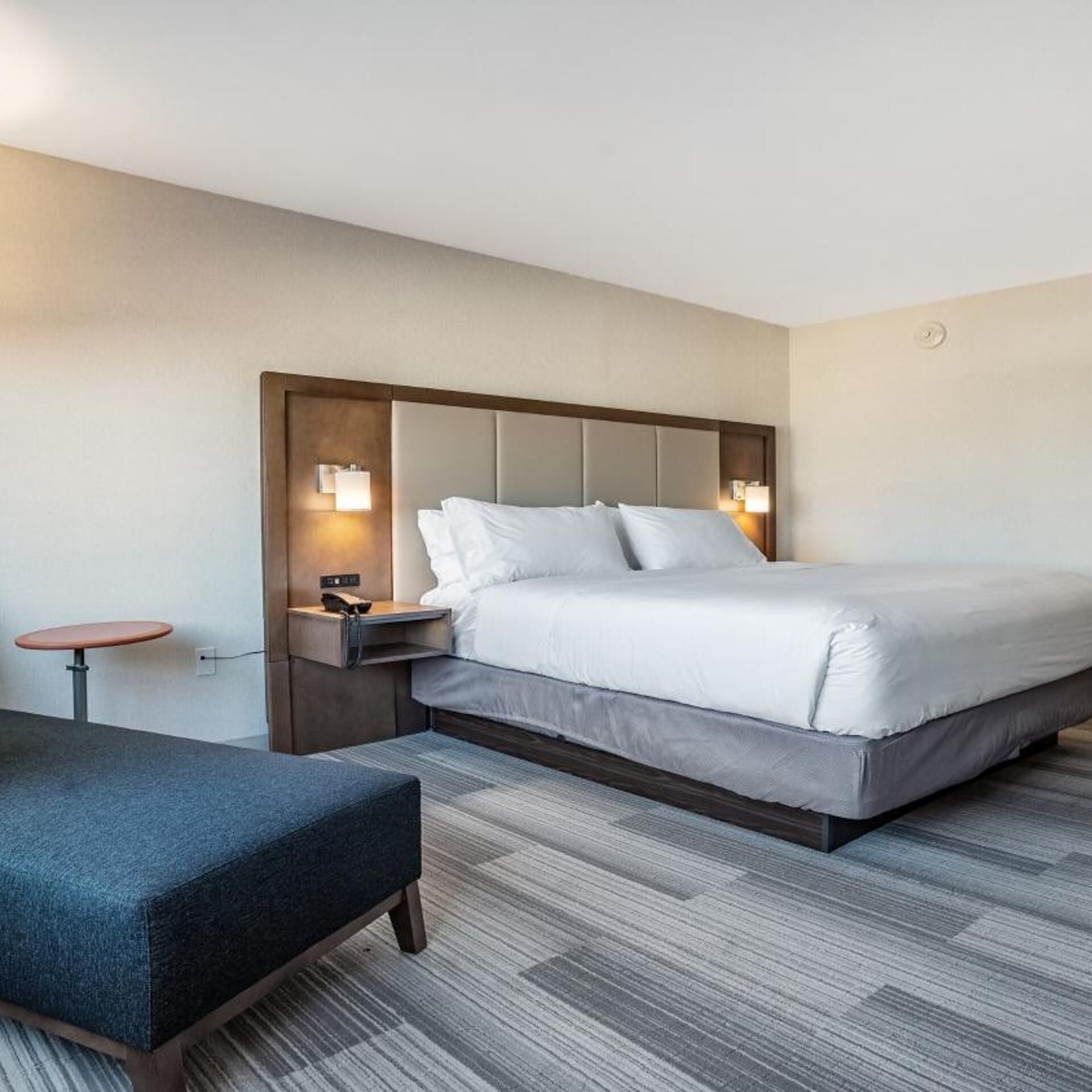 Una habitación de hotel con una cama grande, mesitas de noche, una pequeña mesa redonda y un banco.