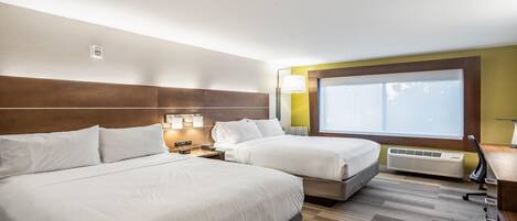 Een hotelkamer met twee bedden, een groot raam met zonwering, een wandheater en een nachtkastje met een lamp.