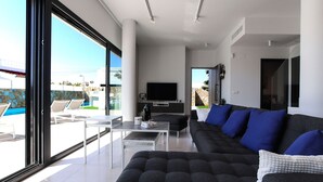 A modern living room with a large window, a flat-screen TV, a sofa with blue cushions, and a pool visible outside.