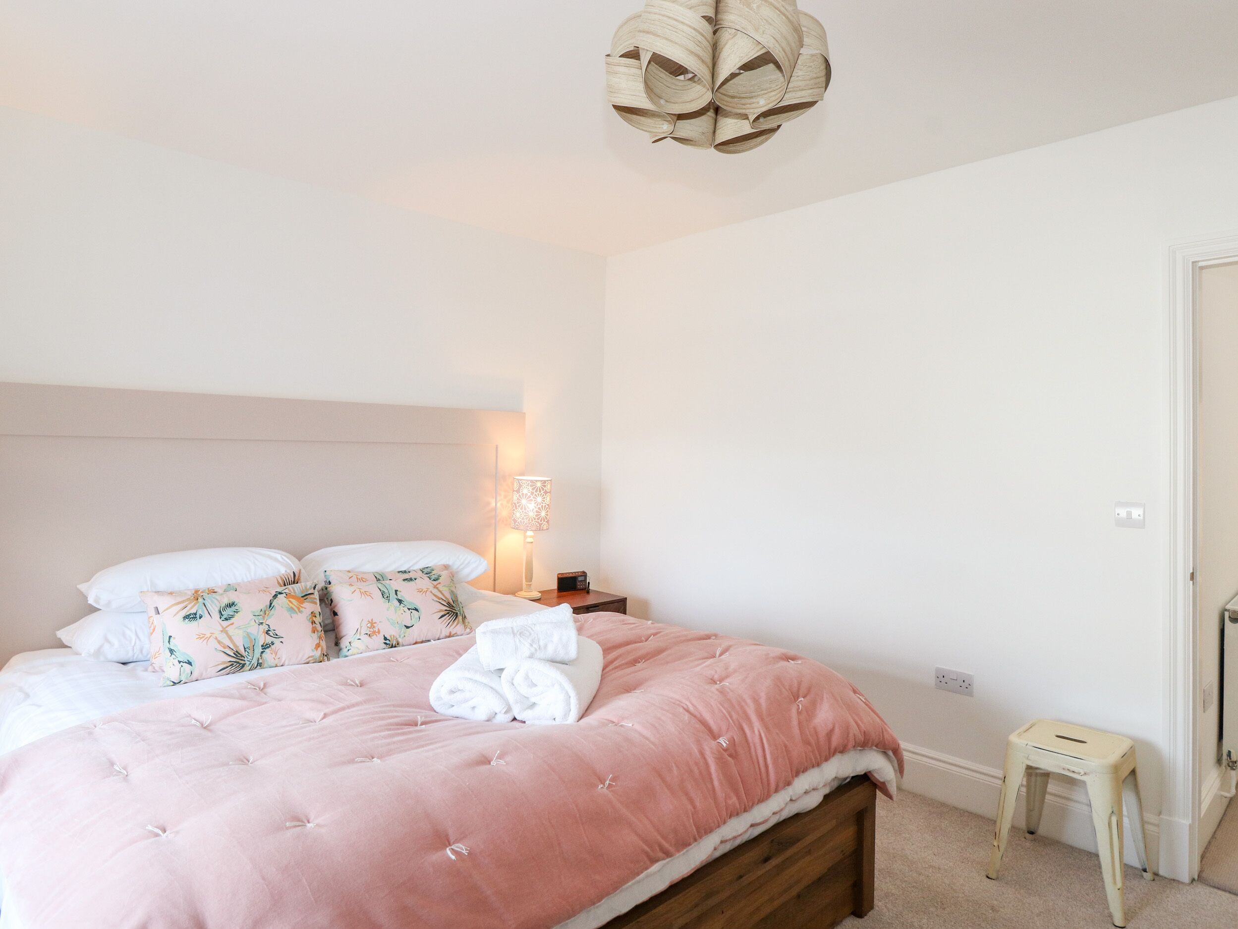 Un lit bien fait, avec une couvre-lit rose et des oreillers blancs, une lampe de chevet et un petit tabouret en bois.