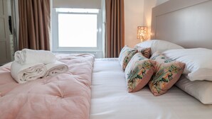 Un lit bien fait, avec une literie rose et des coussins à motifs floraux, une lampe de chevet et une fenêtre avec des rideaux.