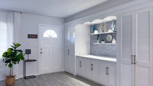 Un couloir avec une porte blanche, une plante en pot et une étagère fixée au mur, ornée d’objets décoratifs.