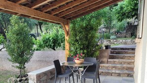 Ein überdachter Patio-Bereich mit Tisch und Stühlen, umgeben von Kübelpflanzen und einer Steinsäule.