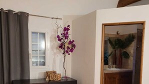 Ein Zimmer mit Holzboden, einem dunklen Holzschrank, einem gewebten Teppich, einer violetten Orchidee und einer Tür zu einem Badezimmer mit Dusche und Waschbecken.
