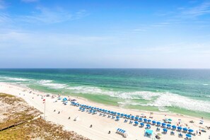 A beach with rows of blue umbrellas and sun loungers, a sandy shore, and the ocean in the background.
