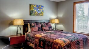 A bedroom with a large bed, two bedside tables, a headboard, a window with blinds, and a painting on the wall.