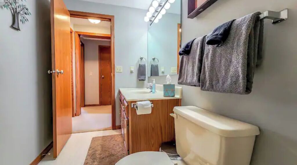 Een badkamer met toilet, wastafel en spiegel. Er is een handdoekrek met handdoeken eraan. Een deur leidt naar een andere kamer.
