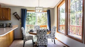 A dining area with a table and chairs, a kitchen counter, and large windows with a view of trees.