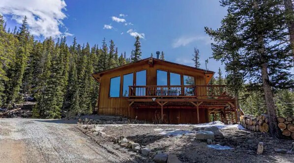A wooden cabin with large windows, a balcony, and a log pile nearby.