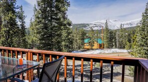 A deck with a glass table and chairs overlooking a snowy mountain landscape and a cabin.