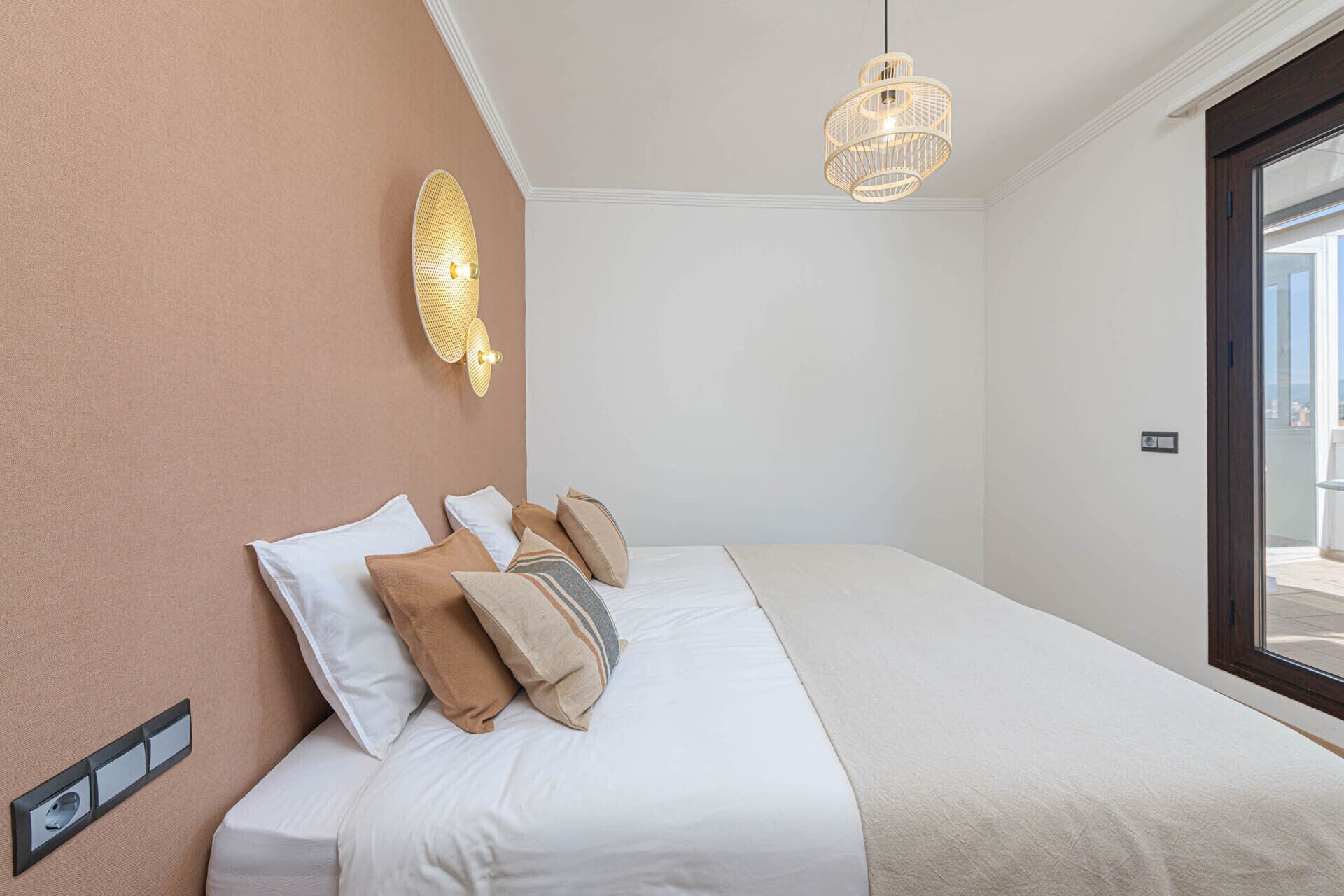Une chambre à coucher avec un lit, des oreillers, un luminaire fixé au mur et une fenêtre.