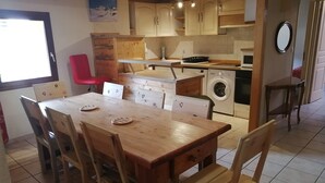 Een kleine keuken met houten kastjes, een eettafel met stoelen, een rode stoel, een wasmachine en een raam.