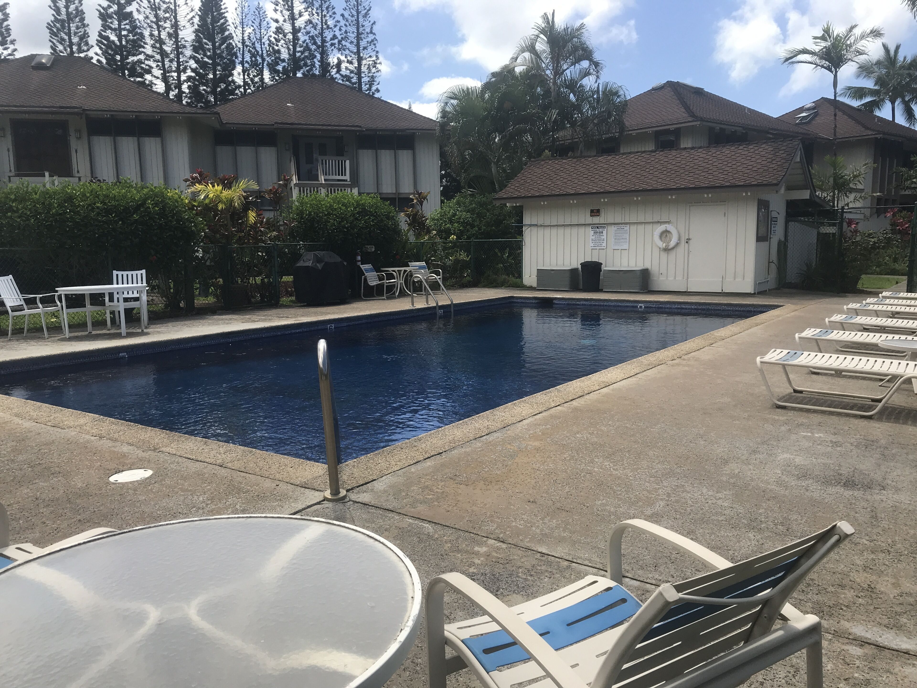 Une piscine entourée de bâtiments et d’arbres, avec des chaises longues et une table à proximité.