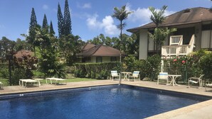 A swimming pool with lounge chairs and a table, surrounded by greenery and a building with a balcony.