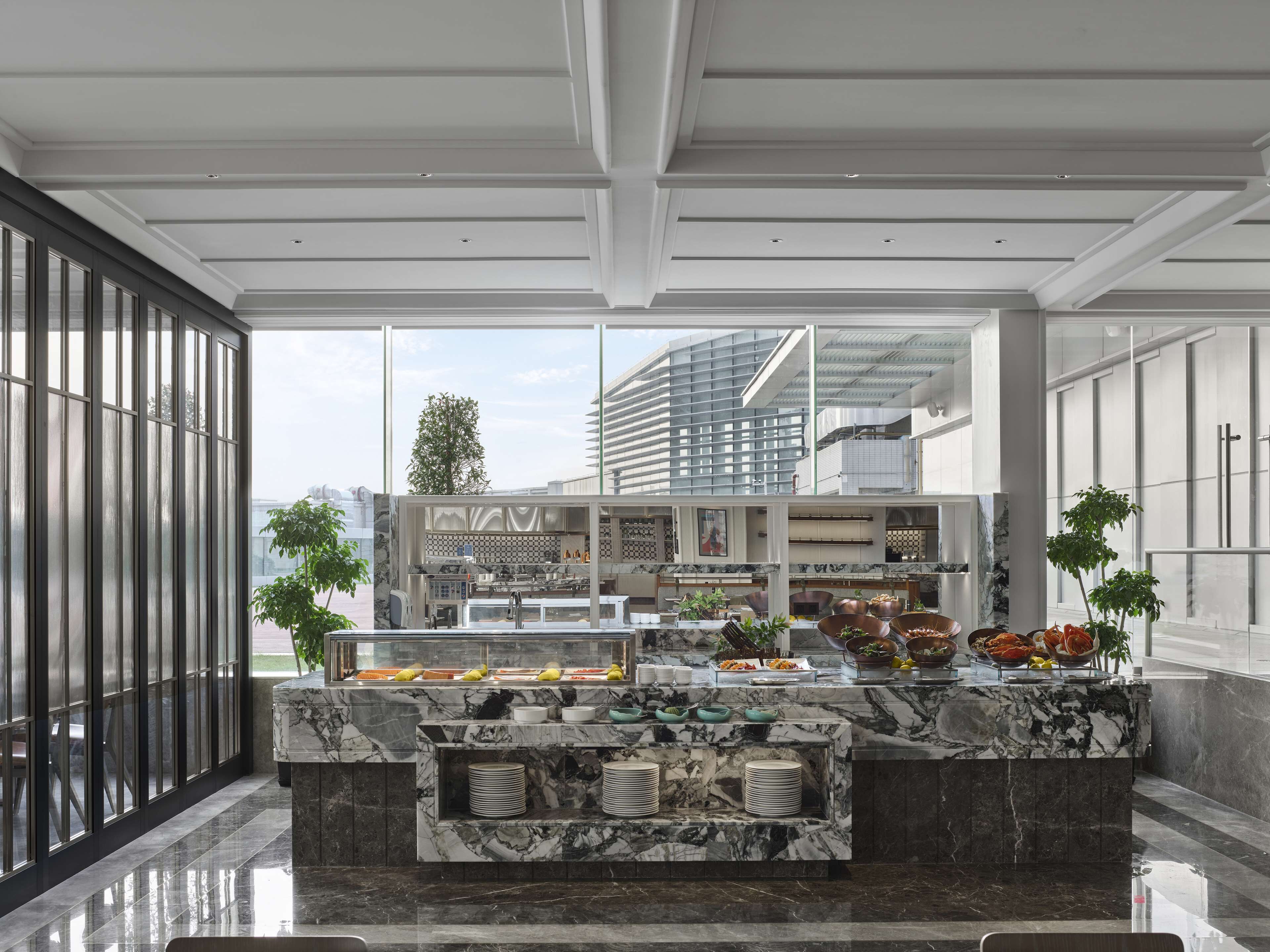 A modern kitchen with marble countertops, a buffet setup, and large windows offering views of the cityscape.
