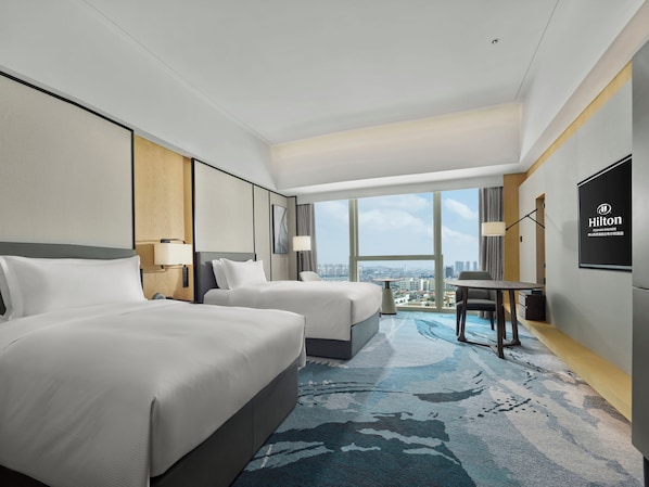A modern hotel room with two beds, a dining table, and a large window offering a city view.