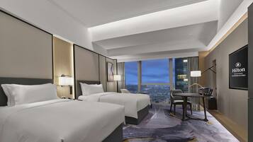 A modern hotel room with two beds, a desk, and a large window offering a city view.