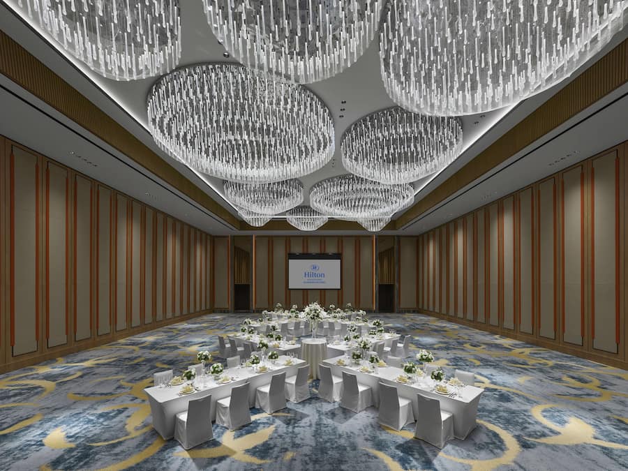 A spacious conference room with a large screen, multiple tables set for dining, and a chandelier with multiple light fixtures.